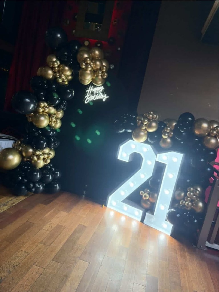 21st birthday setup with black backdrop, black and gold balloons, light-up number sign and Happy Birthday neon